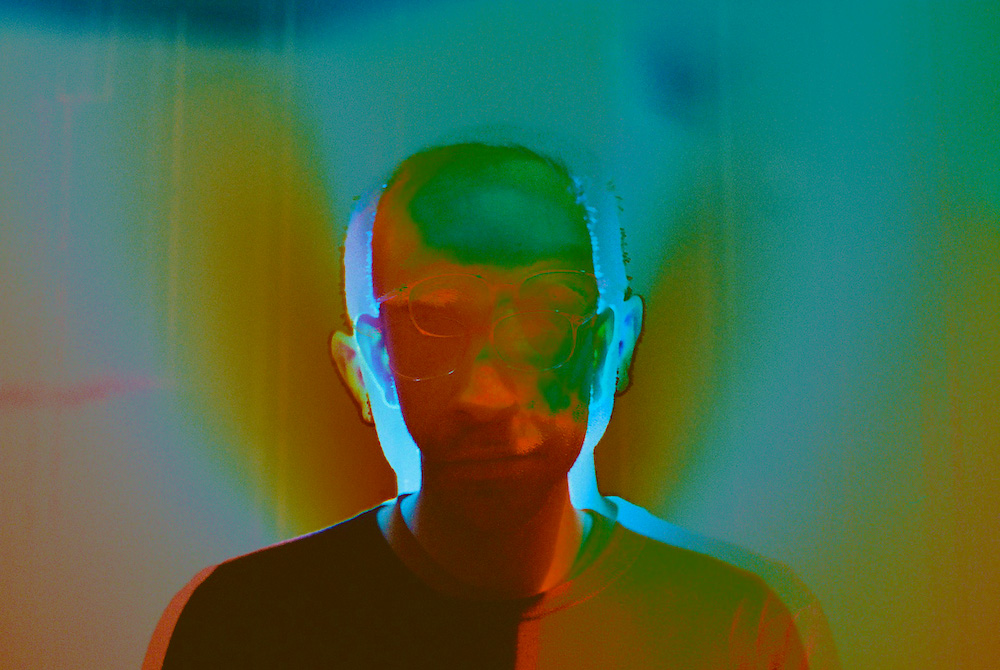 A disorienting composite image of a human being, with overlapping, insdistinct features, made up of multiple exposures of rainbow light, looking in the direction of the camera.